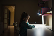 Little Girl Writing In Copybook With Table Lamp In Dark Room