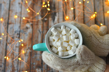 Woman's Hand In White Mittens Holding Mug Of Christmas Cocoa With Marshmallows On Wooden Background With Bokeh New Year's Lights. . Drink For Winter Season. Comfort Food Concept
