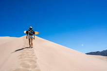 Woman With Sand Board Walking On Edge Of A Sand Dune In The Great Sand Dunes National Park, Colorado United States Of America
