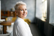 © shurkin_son - Indoor shot of fashionable successful mature blonde woman in formal white blouse turning head to camera with welcoming friendly smile, giving tour around her office. People, age, career and employment