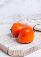 Two ripe persimmon fruits on a wooden board with a towel on a light background