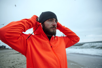 Wall Mural - Outdoor shot of young pretty bearded male with eyebrow piercing holding raised hands behind his head and looking ahead with folded lips, enjoying seaside view after morning workout