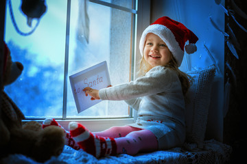 Wall Mural - Cute little girl sitting by the window with a letter to Santa Claus