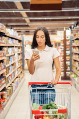 Wall Mural - Daily Shopping. Young woman in the supermarket walking with cart reading article on smartphone concentrated