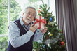 portrait of senior elderly man holding gift box and standing in front of Christmas tree with smile