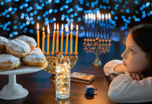 Child Girl Looking At Menorah Candles On Wooden Table And Sufganiyot On Background Light Glitter Bokeh Overlay. Hanukkah Jewish Holiday Israel Hebrew Traditional Family Celebration Invitation Design