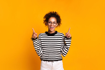 Positive attractive young female with Afro hairstyle, points aside with cheerful expression, shows something and pointing up amazing at blank space,isolated over bright yellow backgroud. Advertisement