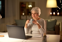 Technology, Old Age And People Concept - Happy Senior Woman With Laptop Drinking Coffee At Home In Evening