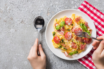 Wall Mural - partial view of woman eating Pappardelle with tomatoes, basil and prosciutto on plaid napkin on grey surface