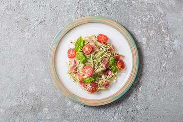 top view of cooked Pappardelle with tomatoes, basil and prosciutto on plate on grey surface