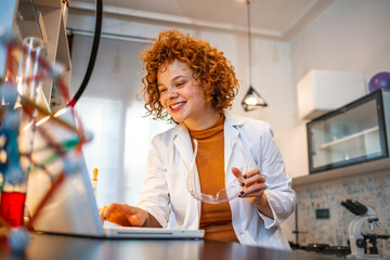 Wall Mural - Working in lab. Portrait of confident female scientist working on laptop in chemical laboratory. Smiling female chemist using laptop for medical research in a laboratory