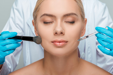 Wall Mural - cropped view of beautician in latex gloves holding cosmetic brush and syringe near attractive girl with closed eyes