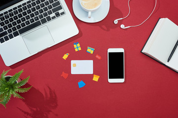 Wall Mural - top view of credit card with icons on red background with smartphone, laptop, earphones, coffee, notebook and plant