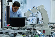 Focus On Robot Arm Which  Picks Up Product From Automated Car On Production Line And An Engineer Working On Laptop At Background. Industry 4.0 Concept; Artificial Intelligence In Smart Factory.