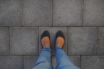 Women Black casual shoes standing and resting on asphalt concrete floor with square tiles. Top View. Concrete floor texture pattern pavement background. Selfie Female of Feet and Legs Seen from Above.