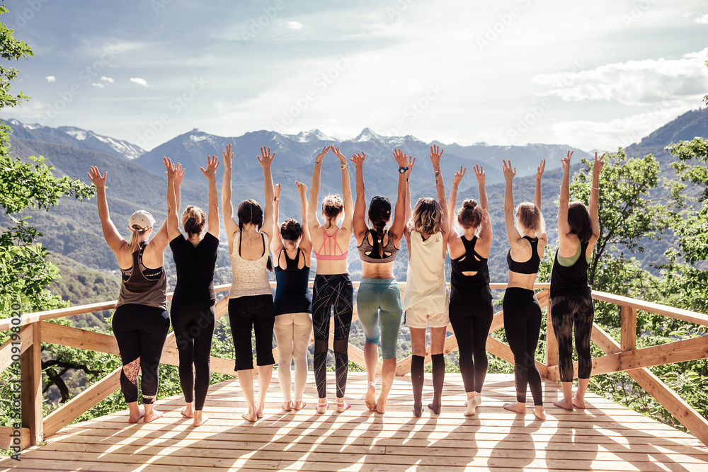 Rear view of group of joyful happy athletic young women in gymnastic clothes raise their hands up and enjoy joint activities in open air among mountains. Concept of sports and personal achievements - obrazy, fototapety, plakaty 