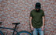 © San4ezz007 - City portrait of handsome hipster guy with beard wearing a blank green military t-shirt and blank black cap standing on a brick wall background. Empty space for your logo or design. Mockup for print.