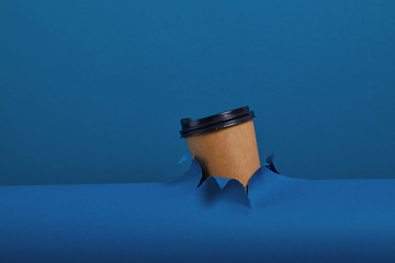 Male hand taking a cup of coffee through torn classic blue paper background. Get energy boost, hot drink in take-away package.