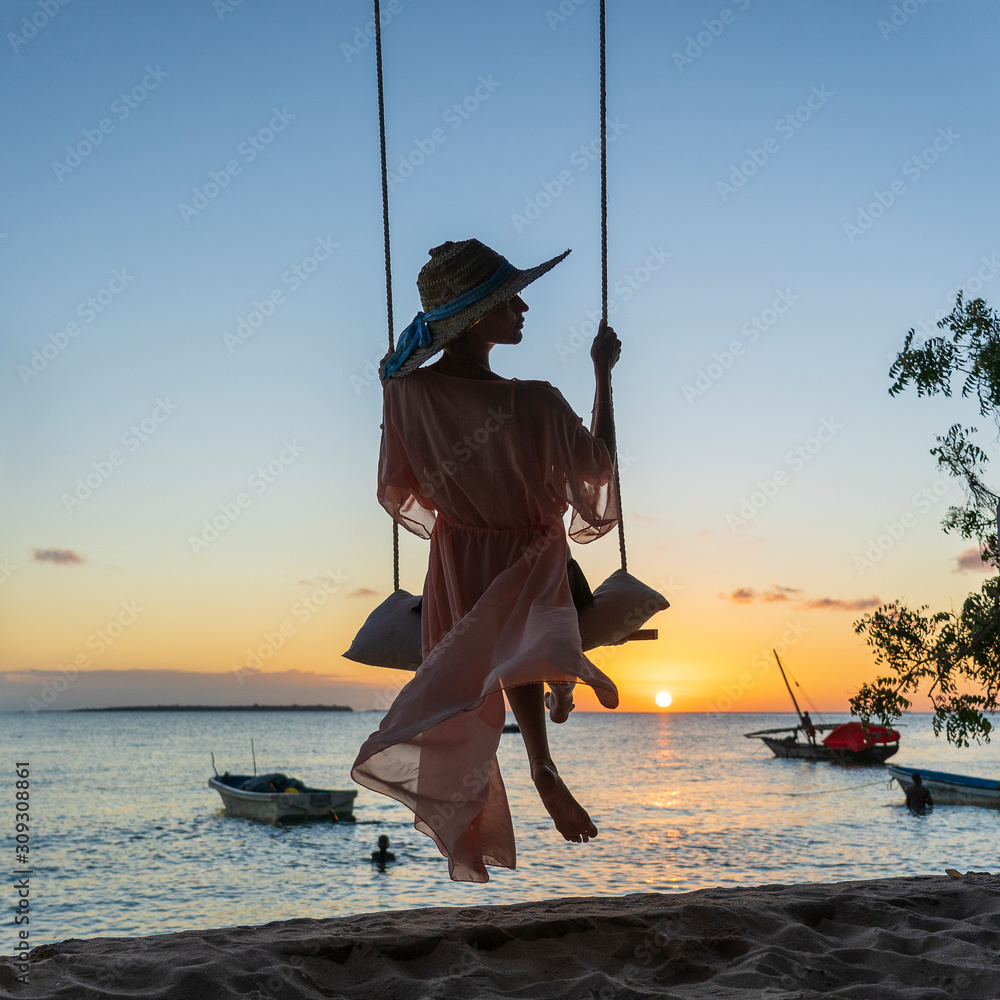 Beautiful girl in a straw hat and pareo swinging on a swing on the beach during sunset of Zanzibar island, Tanzania, Africa. Travel and vacation concept - obrazy, fototapety, plakaty 