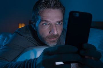 happy man with blue eyes lying on bed late at night in dark light networking on mobile phone or online dating smiling relaxed enjoying internet in social media addiction