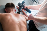 Fototapeta  - Therapist Treating Injury of Professional Athlete Male Patient. Black Massage Gun, Used by Professionals to Massage the Body.Sport Physical Therapy Concept. - Photography