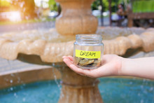 Female Hands Holding A Glass Jar With Coins, Dream Note Yellow Sticker. Saving Of Money For Trips, Travel. Financial Pillow. The Concept A Deposit For Your Dreams. On The Background Of The Fountain