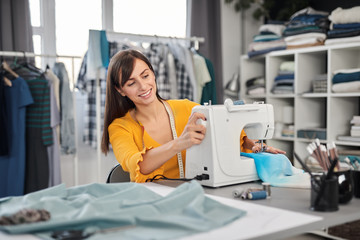 Smiling charming caucasian fashion designer sitting in her studio and sewing beautiful evening dress.