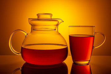 Glass teapot with a filled glass, on a yellow gradienton dark background.