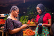 © Confidence - African woman shopping food stuff in a local market paying by doing mobile transfer via phone for a trader