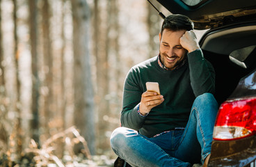 Handsome young man using his smart-phone and smiling while he's sitting in open car trunk in the forest.