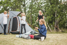 Teenage Girl Using Smartphone While Family Pitching Tent At Campsite