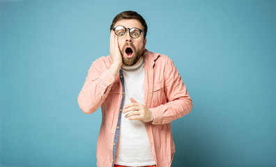 Man in shifted glasses is shocked, he opened mouth, looks at the camera and holds hand to face. Isolated on a blue background.