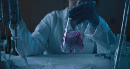 female research scientist mixing up a conical flask.blue lighting in a dark lab room.medium, dolly o