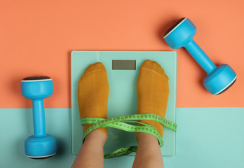 Fitness, slimming concept. Woman measures her weight by standing her feet in socks on scale against pink blue background with dumbbells. Top view