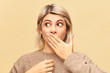 Leinwandbild Motiv Genuine human emotions, feelings and reaction. Attractive blonde young woman having surprised look, trying to keep secret or confidential information, covering mouth with hand and looking away