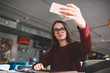 © GalakticDreamer - Trendy female blogger holding mobile phone in hand looking at cellphone camera and taking photo of herself, caucasian hipster girl sitting in favourite cafe and using smartphone clicking selfie photo