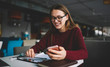 © GalakticDreamer - Smiling student girl in eyeglasses sitting in loft interior cafe laughing reading news on website via cellphone, positive funny woman chatting with friends online on modern smartphone while learning