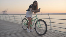 Young Attractive Woman Uses A Smartphone And Riding Vintage Bike Near The Sea During Sunrise Or Sunset