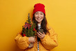 © WHstudio Leushin N - Charming tender Korean woman with no makeup, toothy smile, holds small decorated Christmas firtree, uses mobile phone for sending text messages, dressed in winter outerwear, gets notification.
