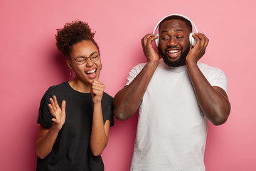 Wall Mural - Joyful curly haired woman pretends singing in microphone plump bearded man listens music in headphones, have broad smiles, enjoy favourite playlist, relax after hard working week. People and rest