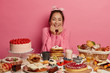 Feminine girl smiles pleasantly, keeps both hands under chin, dressed in knitted rosy sweater, poses at table with sugary desserts and bakery, thinks what to eat first, isolated over pink background