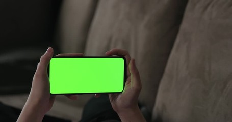 Young woman lying on a couch and using smartphone with horizontal green screen