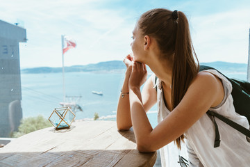 a tourist with a ponytail and a backpack behind her sits at a wooden table and looks at the sea