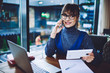 © GalakticDreamer - Positive hipster girl in optical spectacles talking with friend via application on modern cellphone while sitting at cafeteria table with laptop computer and digital gadget for planning organisation