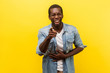 Portrait of joyful positive man in denim shirt with rolled up sleeves holding hand on belly and laughing out loud, pointing at camera, mocking you. indoor studio shot isolated on yellow background