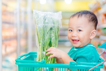 Wall Mural - Funny little asian boy child smile and laughing he choosing vegetable fist in hypermarket, Little asian toddler boy sitting in the trolley cart during family shopping in supermarket.