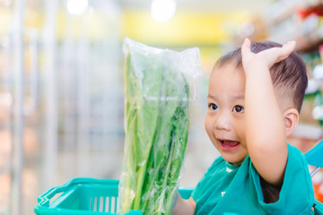 Wall Mural - Funny little asian boy child smile and laughing he choosing vegetable fist in hypermarket, Little asian toddler boy sitting in the trolley cart during family shopping in supermarket.