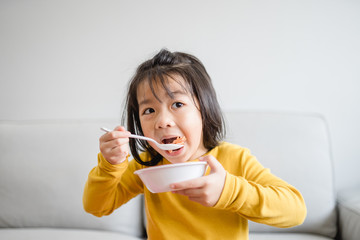 Wall Mural - Hungry little asian girl eating rice with salmon teriyaki and watching TV in sofa at home.Happy meal and Delicious asian food concept.