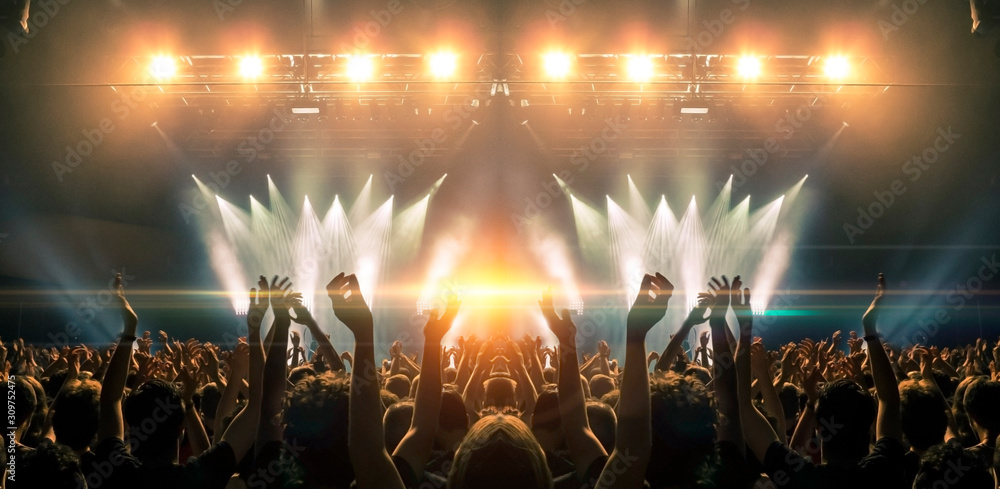 Photo of a concert hall with people silhouettes clapping in front of a big stage lit by spotlights. Shot is taken from concert crowd point of view, lens flare is visible. - obrazy, fototapety, plakaty 