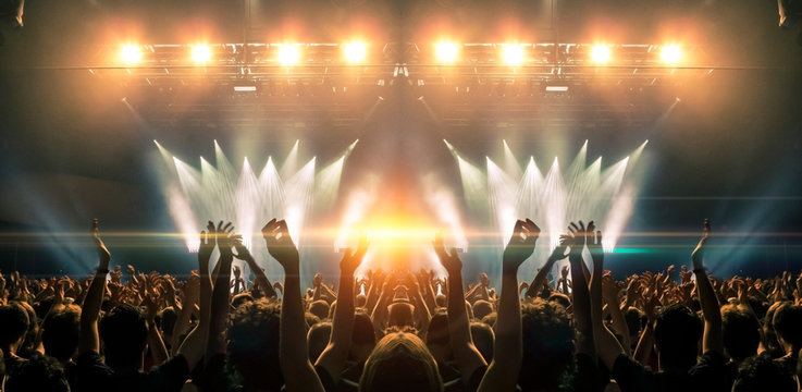 photo of a concert hall with people silhouettes clapping in front of a big stage lit by spotlights. 